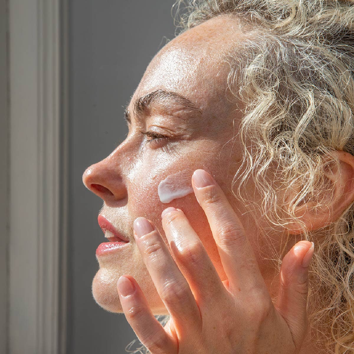 Woman applying hyaluronic acid moisturizing cream to her face with a neutral background